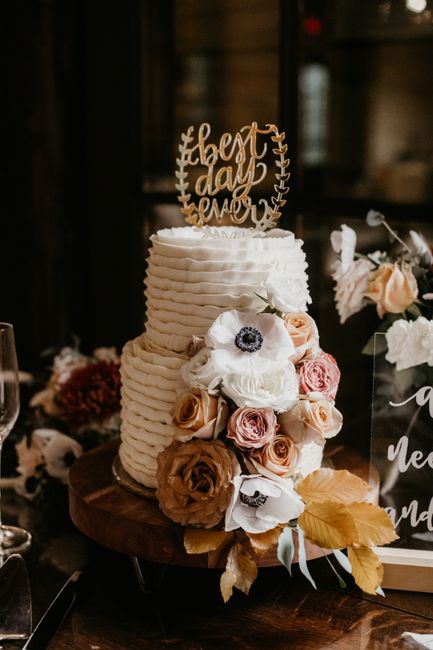 A three-tiered white wedding cake with textured, wavy icing decoration, topped with a wooden cake topper reading 'best day ever' in cursive script. The cake is adorned with a cascading arrangement of