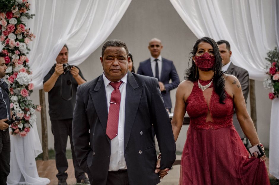 A woman dressed in a burgundy lace sleeveless dress and matching face mask walks hand-in-hand with a man in a dark suit and burgundy tie, during a wedding ceremony. They are outdoors under a white dra