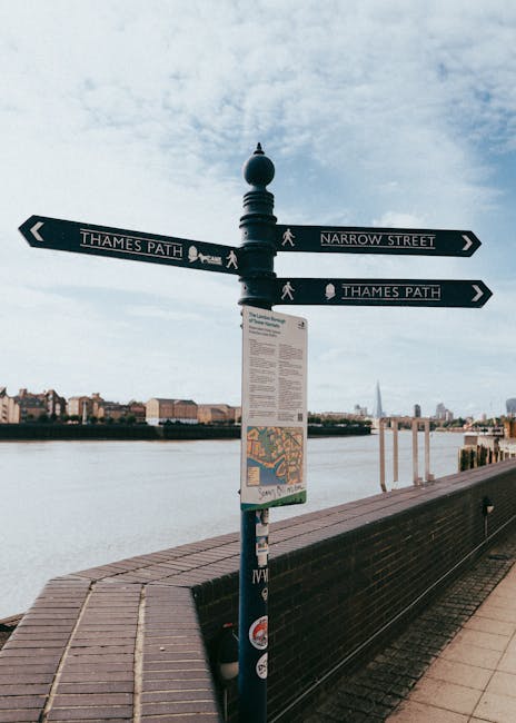 A close-up of a black signpost on a riverside promenade, featuring four directional arrows labeled 'Thames Path' and 'Narrow Street', with white walking man icons, mounted on a decorative black finial